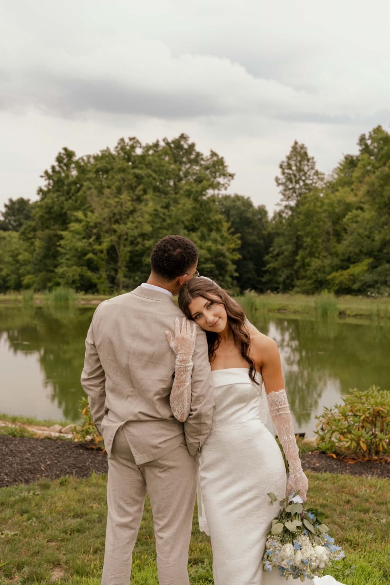 Bride leaning on her groom's shoulder holding a bouquet, wearing a strapless gown and lace gloves by a pond