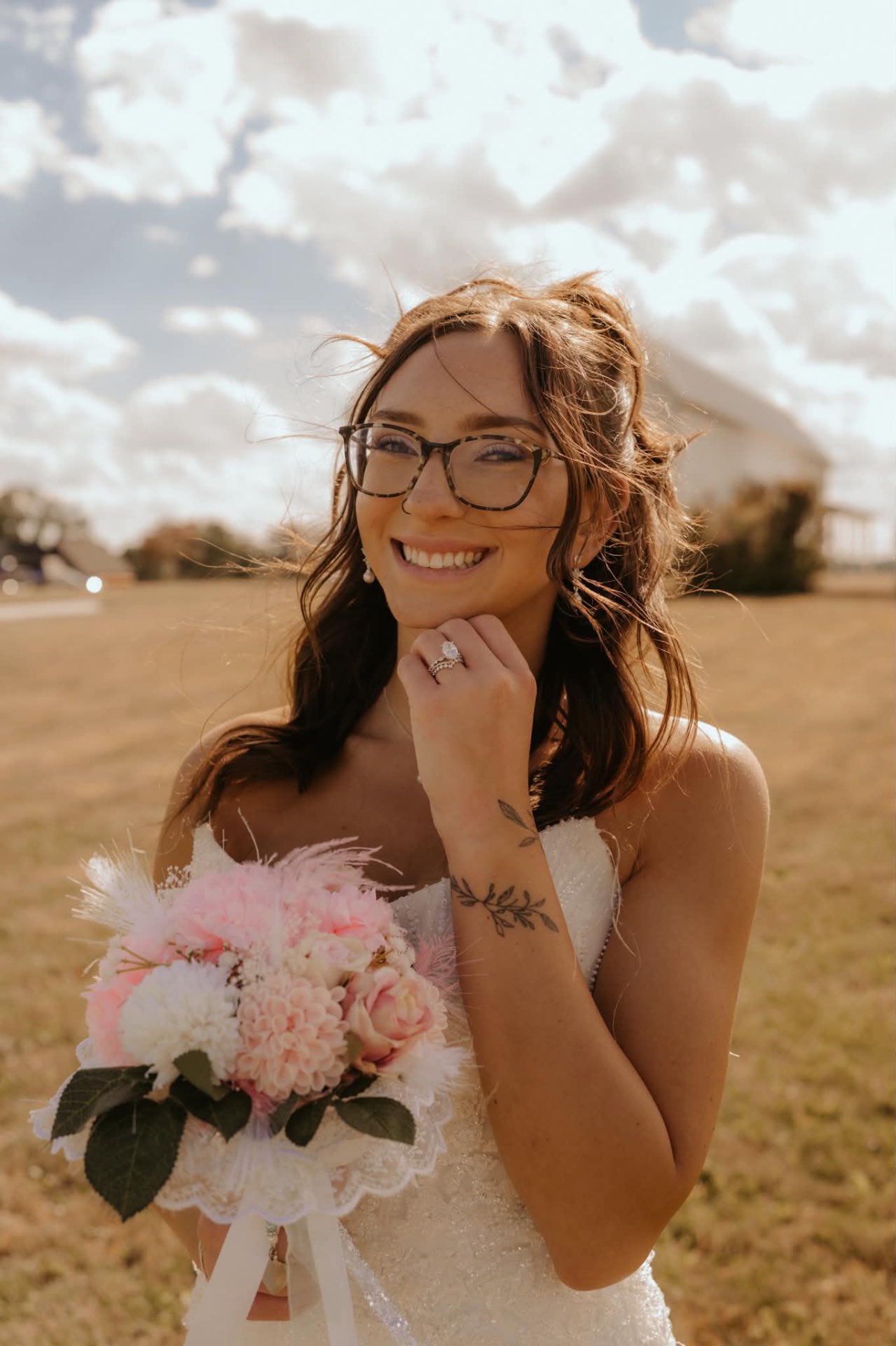 Bride in a strapless lace gown and glasses smiling in a sunny field, holding a pink and white bouquet with soft glam bridal makeup