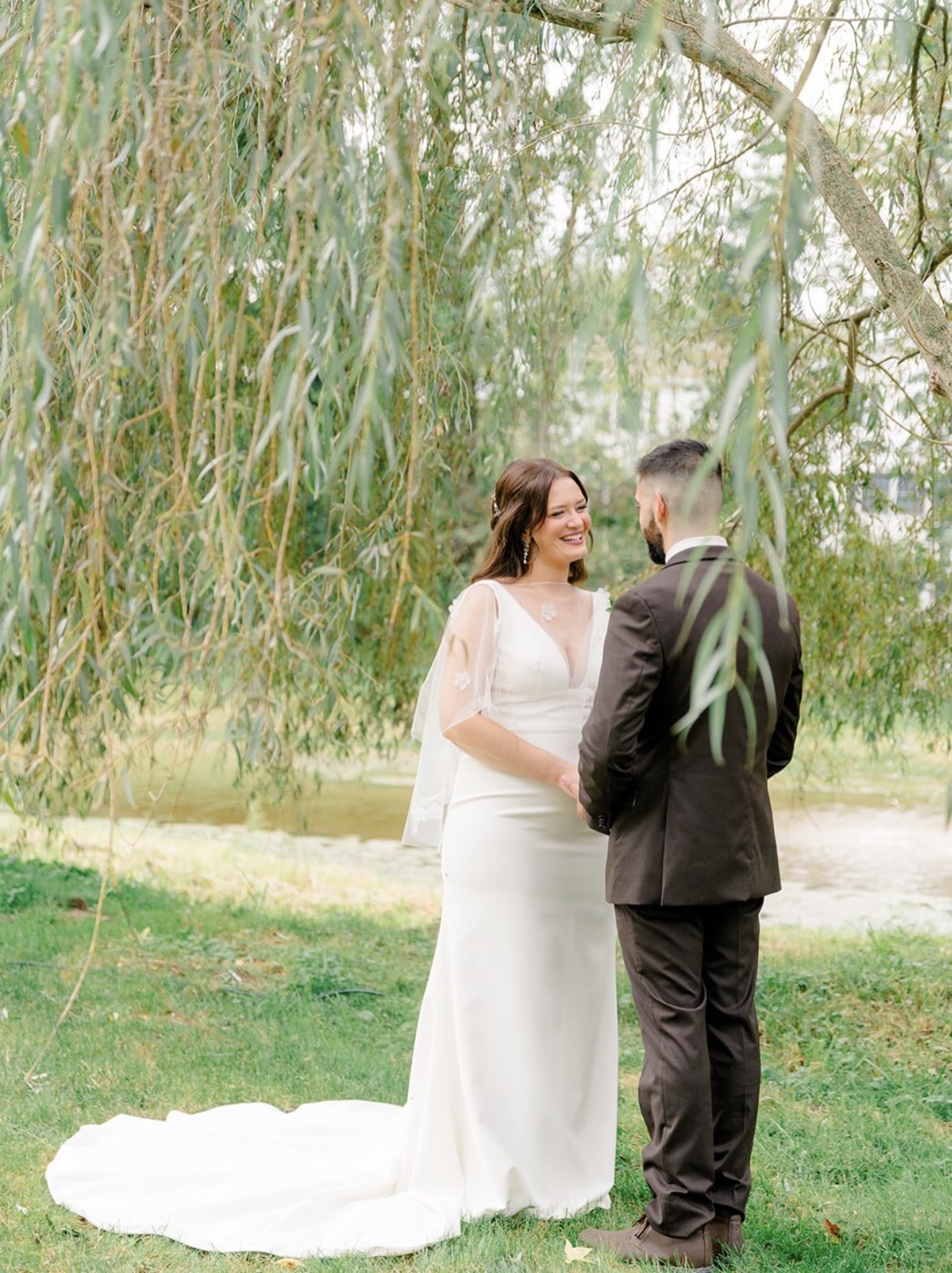 Bride with soft bridal makeup and a groom in a brown suit holding hands under a weeping willow tree