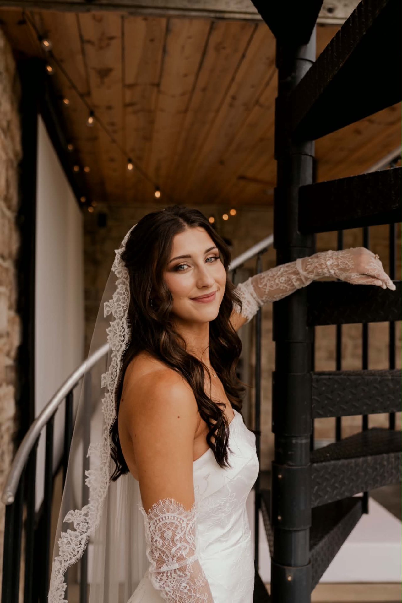 Bride in a strapless gown and lace gloves smiling beside a spiral staircase with soft glam bridal makeup