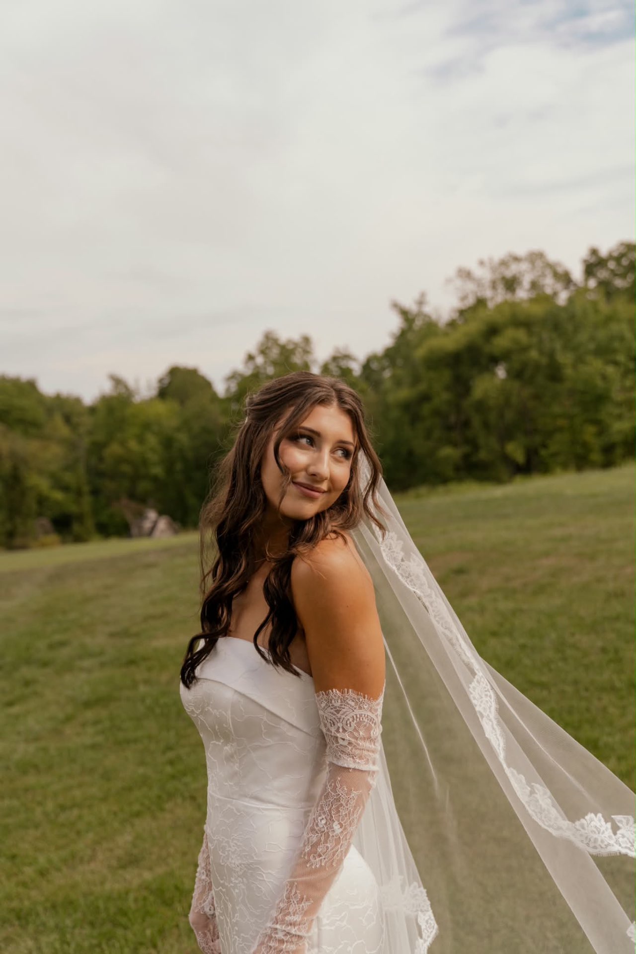 Bride in a strapless floral brocade gown and lace veil looking over her shoulder in a grassy field, soft glam bridal makeup