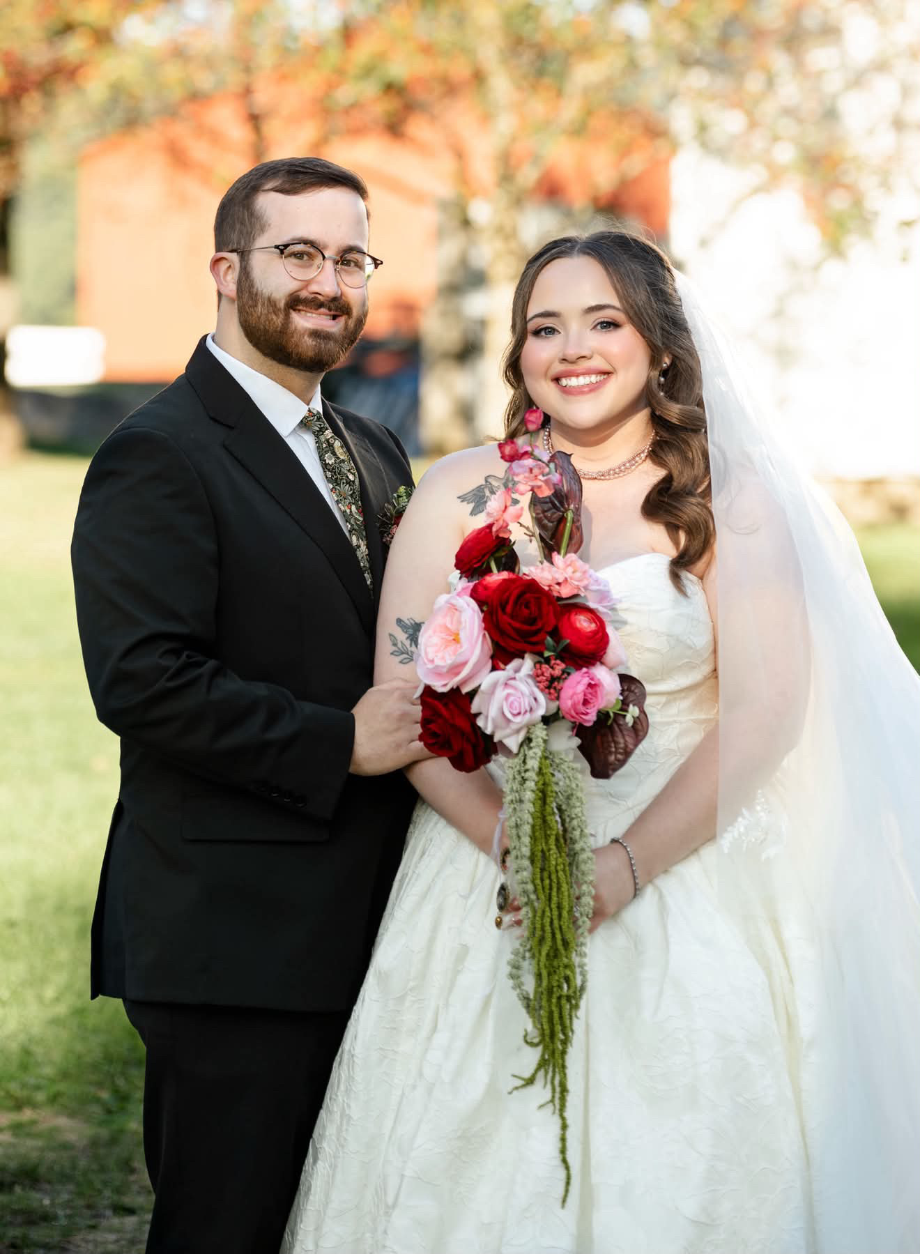 Bride and groom standing together on a lawn, bride in a strapless ivory ballgown and veil holding a deep red and blush cascading bouquet