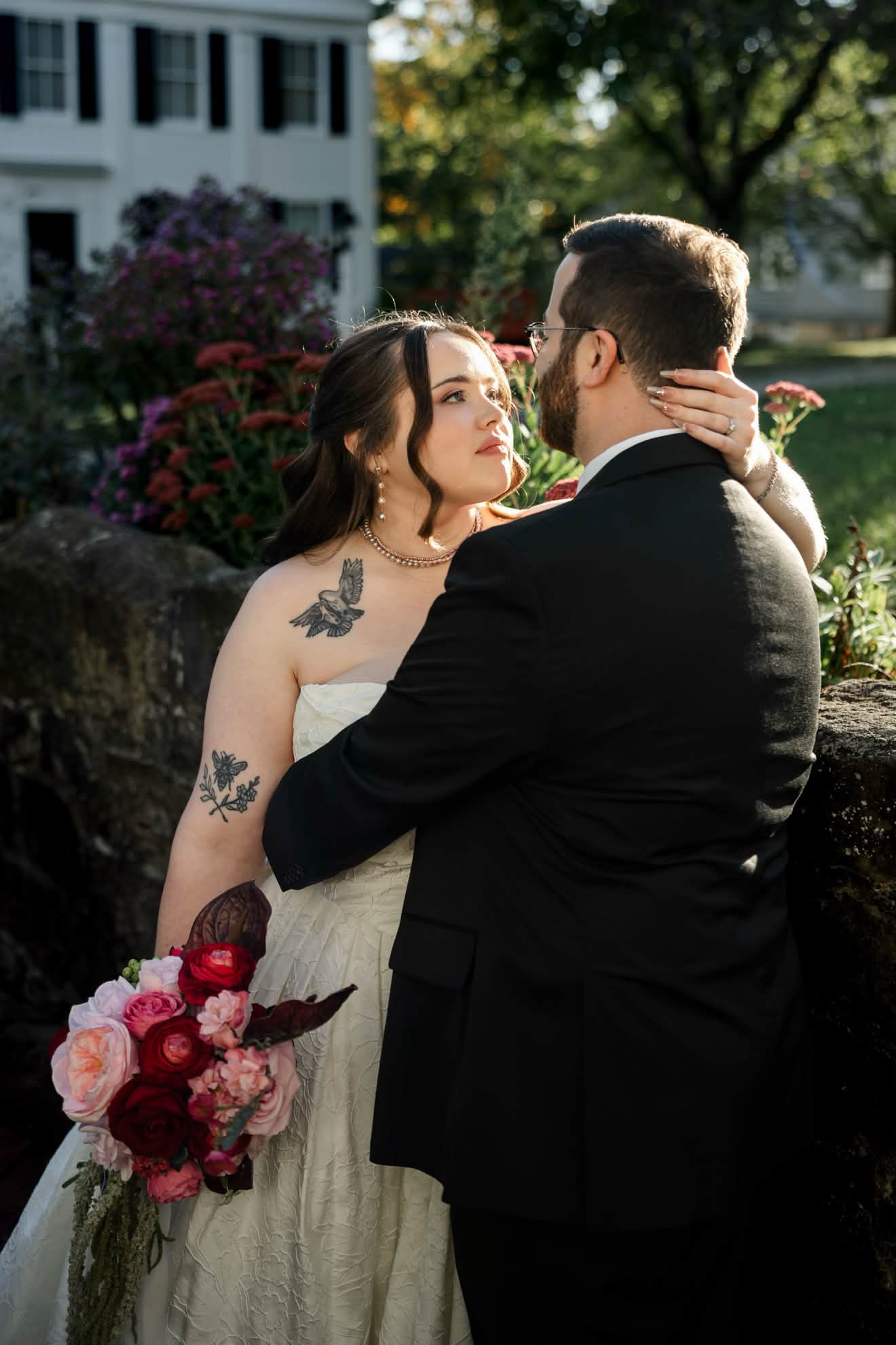 Bride and groom in a close embrace in a golden-hour garden, bride in a strapless damask gown holding a crimson and blush bouquet