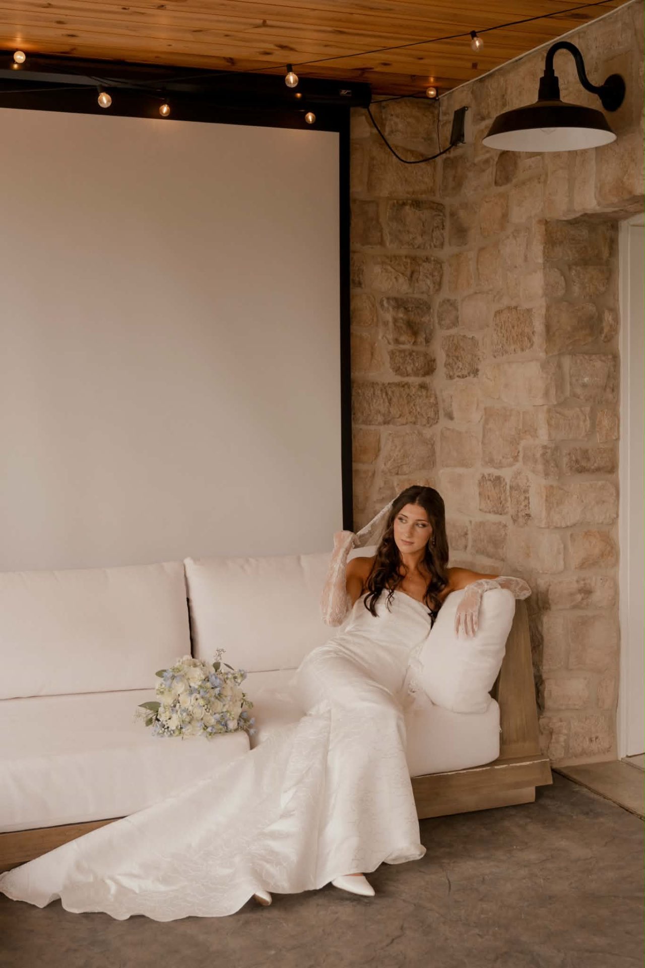Bride lounging on a white sofa in her floral brocade gown and veil with a bouquet, soft glam bridal makeup