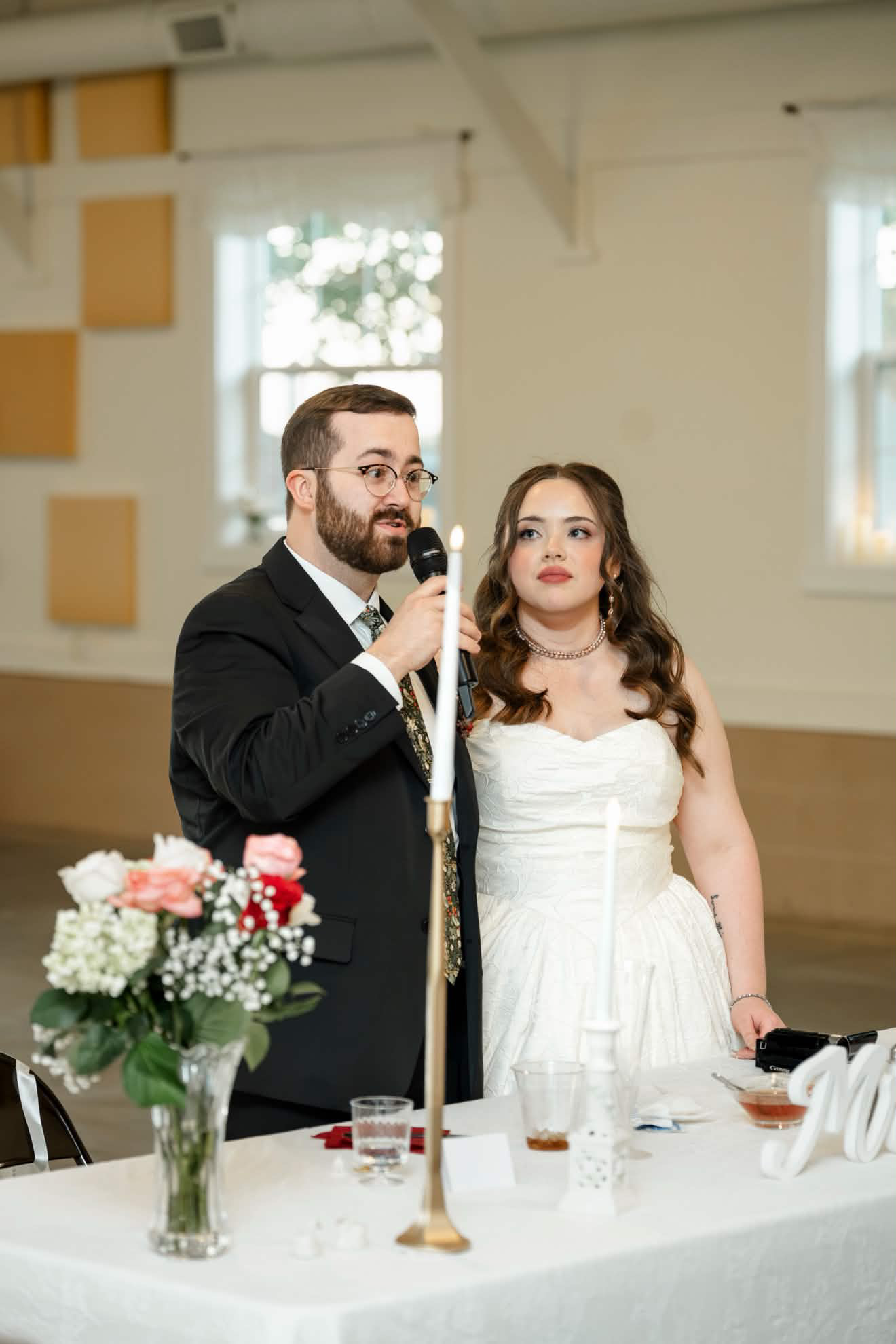 Bride and groom behind the reception head table during a toast, bride in a strapless gown and pearl choker with soft glam bridal makeup