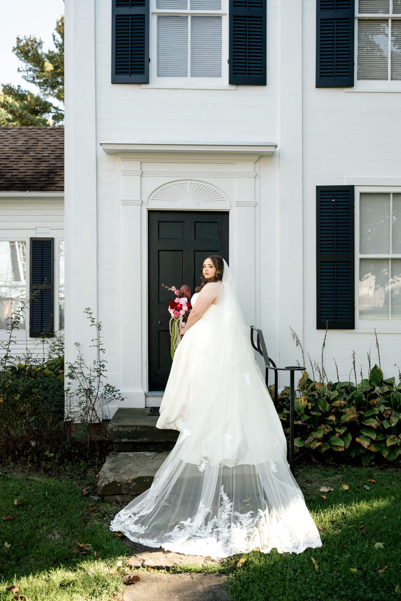 Bride in a strapless ivory gown and embroidered cathedral veil standing on stone steps, looking back over her shoulder with a bouquet
